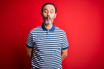 Middle age hoary man wearing casual striped polo standing over isolated red background making fish face with lips, crazy and comical gesture. Funny expression.