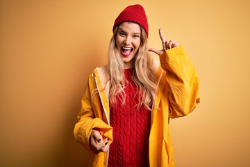 Young beautiful blonde woman wearing raincoat and wool cap over isolated yellow background pointing finger up with successful idea. Exited and happy. Number one.