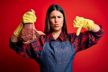 Young butcher woman holding fresh raw beef meat stake over red background with angry face, negative...