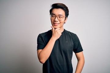 Young handsome chinese man wearing black t-shirt and glasses over white background with hand on chin thinking about question, pensive expression. Smiling with thoughtful face. Doubt concept.
