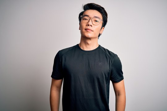 Young handsome chinese man wearing black t-shirt and glasses over white background Relaxed with serious expression on face. Simple and natural looking at the camera.