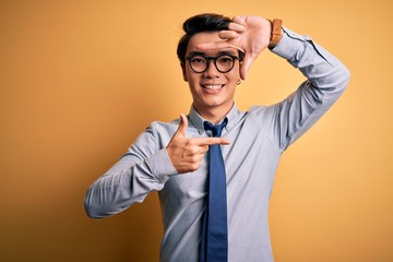Young handsome chinese businessman wearing glasses and tie over yellow background smiling making frame with hands and fingers with happy face. Creativity and photography concept.