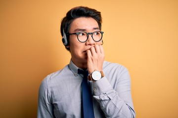 Young handsome chinese call center agent man wering glasses using headset looking stressed and nervous with hands on mouth biting nails. Anxiety problem.