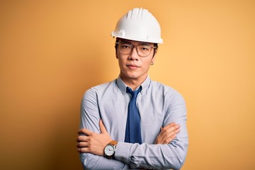 Young handsome chinese architect man wearing safety helmet and tie over yellow background skeptic and nervous, disapproving expression on face with crossed arms. Negative person.