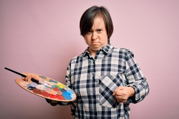 Young down syndrome artist woman holding painter palette and paintbrush over pink background...