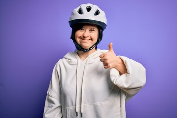 Young down syndrome cyclist woman wearing security bike helmet over purple background doing happy thumbs up gesture with hand. Approving expression looking at the camera showing success.
