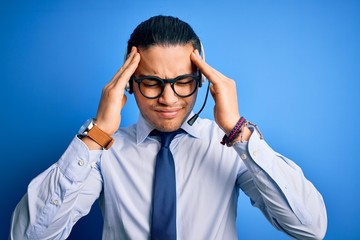 Young brazilian call center agent man wearing glasses and tie working using headset suffering from headache desperate and stressed because pain and migraine. Hands on head.