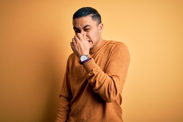 Young brazilian man wearing casual sweater standing over isolated yellow background smelling something stinky and disgusting, intolerable smell, holding breath with fingers on nose. Bad smell