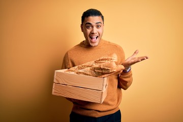 Young baker brazilian man holding box with homemade bread over isolated yellow background very...