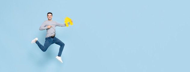 Happy handsome man jumping and pointing to yellow house model isolated on blue banner background...