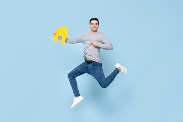 Happy handsome man jumping and pointing to yellow house model isolated on blue studio background