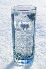 glass full to the brim of fresh crystal clear transparent water with frozen splash from thrown snowball on the white cold snow
