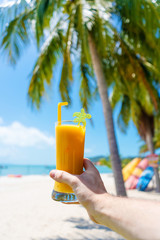 First-person view. Girl holds a glass cup of cold mango fresh on the background of a sandy tropical beach. White sand and palm trees. Fairytale vacation