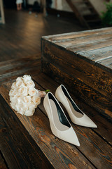 Elegant stylish white wedding shoes with a pointed toe on a long transparent heel and a wedding bouquet on a dark wood staircase background. Vertical