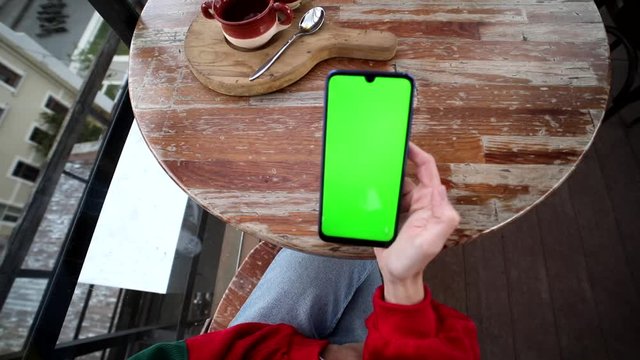 Top of young beautiful woman hands holding smartphone with green screen sitting in modern cafe outdoors, drinking coffee. Chroma-key display, mockup, free app, online application. Round table.