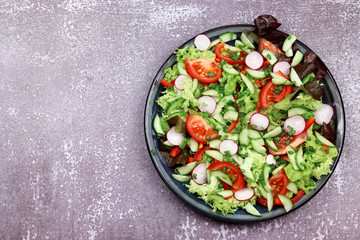 Leaf lettuce with cherry tomatoes, green onions, garlic, bell pepper and radish in a dark plate on a dark wooden background. Top view, flat lay
