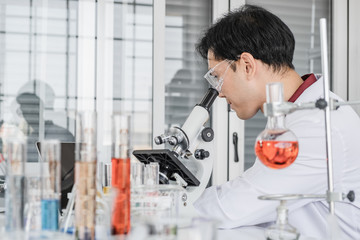 A male scientist with black hair wearing white coat and protective glassware working on laptop in a laboratory setting with test tubes and microscope.