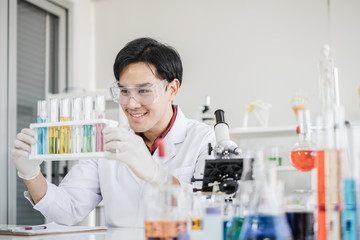 A male scientist with black hair wearing white coat and protective glassware sitting smiling looking at test tube and solutions in white laboratory room.