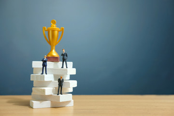 Miniature business concept - businessman standing on white staircase ladder with golden trophy...
