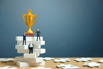 Miniature business concept - businessman standing on white staircase ladder with golden trophy...