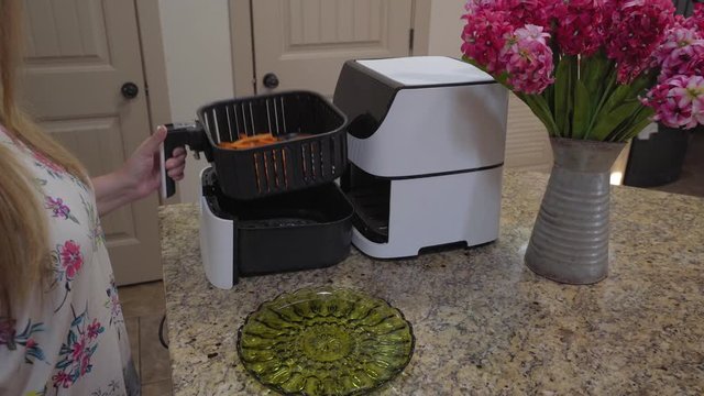 A woman pulls out a batch of freshly cooked sweet potato fries from an electric air fryer and empties it onto a plate, static medium shot