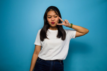 Fototapeta premium Portrait of Young beautiful asian women using white T-shirt with blue isolated background, showing peace sign and smiling, Victory sign, two sign