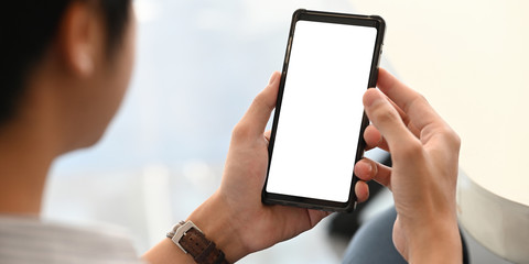 Photo of creative man holding a cropped black smartphone with white blank screen in hands while sitting and relaxing at home with blurred living room as background.