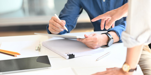 Cropped image of architect man's hands holding a stylus pen and computer tablet for drawing a project concept with colleague at the modern working desk with accessories putting on it.