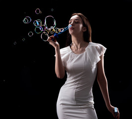 A beautiful slender girl in a white dress lets soap bubbles on a black background, adults are...
