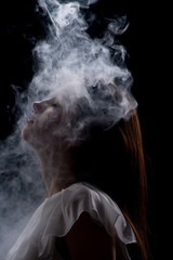 The silhouette of a young beautiful girl in a white dress in a cloud of smoke, a cloud of thick smoke descends on the woman 's face