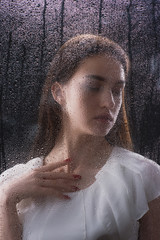 A beautiful slender brunette with a good figure behind a glass with raindrops, the silhouette of a woman behind a blind glass on a dark background