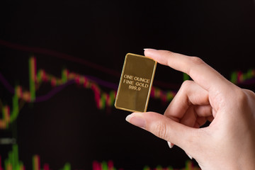 The hand holds a gold bar on the background of a laptop on which there is a trading price chart. One ounce of gold in hand. Trading gold on the stock exchange and Forex.