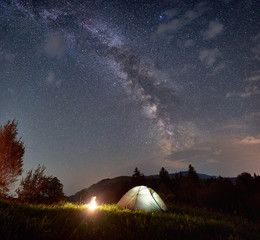 Fototapeta na wymiar Night camping in the mountains at summer. Illuminated tourist tent and bonfire under incredible evening sky full of stars and Milky way. Beauty of the nature