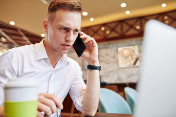 Frowning young businessman reading data on laptop screen and talking on phone with client or coworker