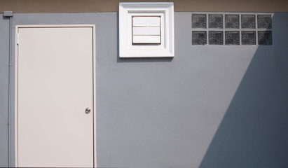 Front view of white door, square ventilation shutter and glass blocks with sunlight and shade on surface of gray cement wall with brown stripe, image for home exterior decoration design concept
