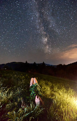 Vertical snapshot of a girl lying in summer grass at night observing beautiful sky full of stars and Milky way in the mountains. Concept of unity with nature