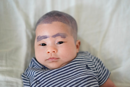 Close up one month old baby apply Butterfly pea flower to eyebrows and hair, in Thai called Anchan,baby lay down on white bed sheet background.