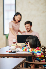 Fototapeta premium Creative talented little girl painting on paper with gouache, her parents talking in background