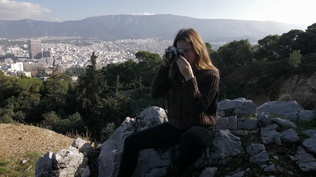 Young woman sitting on rock in Lycabettus hill takes photo with her analog camera in Athens, Greece