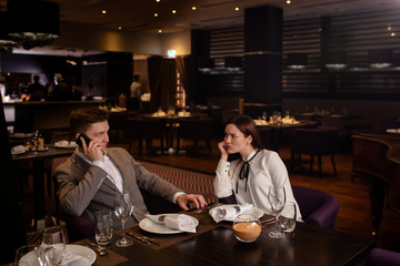 offended caucasian woman and handsome guy sit in restaurant, they don't talk with each other. beautiful couple came to celebrate anniversary, but they had quarrel. man talk on phone