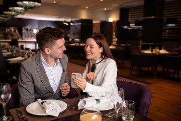 young pretty lady happily telling interesting story to her boyfriend while they have dinner, they are on date, she show on smartphone and laugh