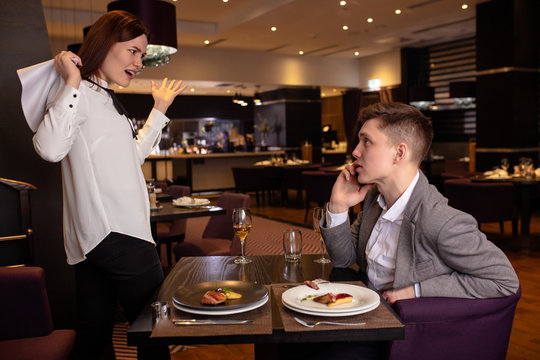 Caucasian Couple Quarreling In Restaurant. Woman Yell At Man Talking On Phone. Lady Stand In A Fury, Want To Go Away From Him