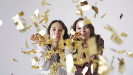 Two beautiful girls blowing gold glitter confetti on a white background