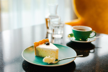 A slice of cheesecake on a blue plate, dessert fork with a small piece of pie, a cup of black coffee, a carafe of water and a glass, on a black table with reflections. Back natural light, soft focus