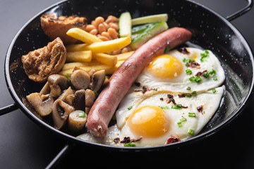 Traditional English breakfast with egg, mushrooms, cucumber and beans on a black pan background.