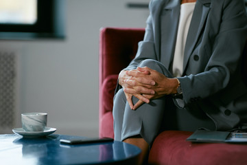 Close up of unrecognizable businesswoman waiting in cafe, focus on hands clasped over knee and coffee cup, copy space