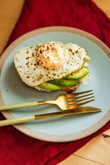 A rye-bread toast with cheese, avocado and fried egg for breakfast, served on a blue plate. Beautiful golden cutlery, red napkin.