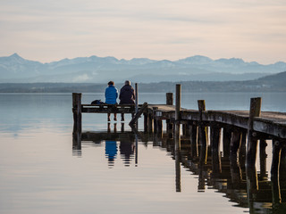 Rear view of couple sitting on wooden pier at lake ammer in bavaria with alps in the background