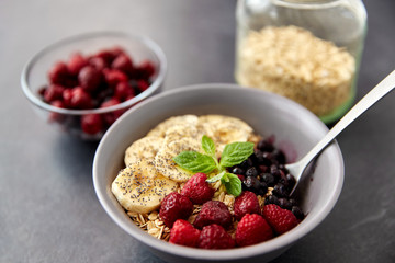 food and breakfast concept - oatmeal cereals in bowl with wild berries, banana and spoon on slate stone table