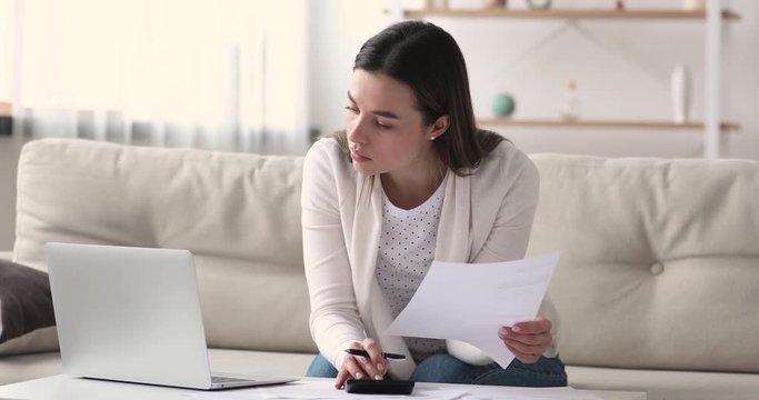 Young adult woman renter checking household payments using calculator and laptop sitting on sofa. Serious lady calculating bills, managing finances, paying rent or bank loan, counting taxes at home.